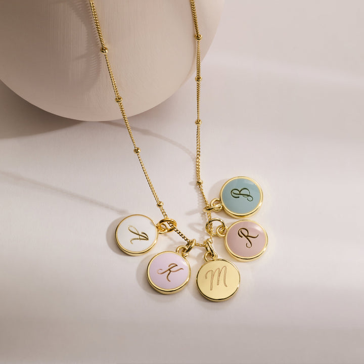 Initial charm necklace with five round enamel discs in pastel colors, each engraved with a script initial, on a chain with a ring clasp.