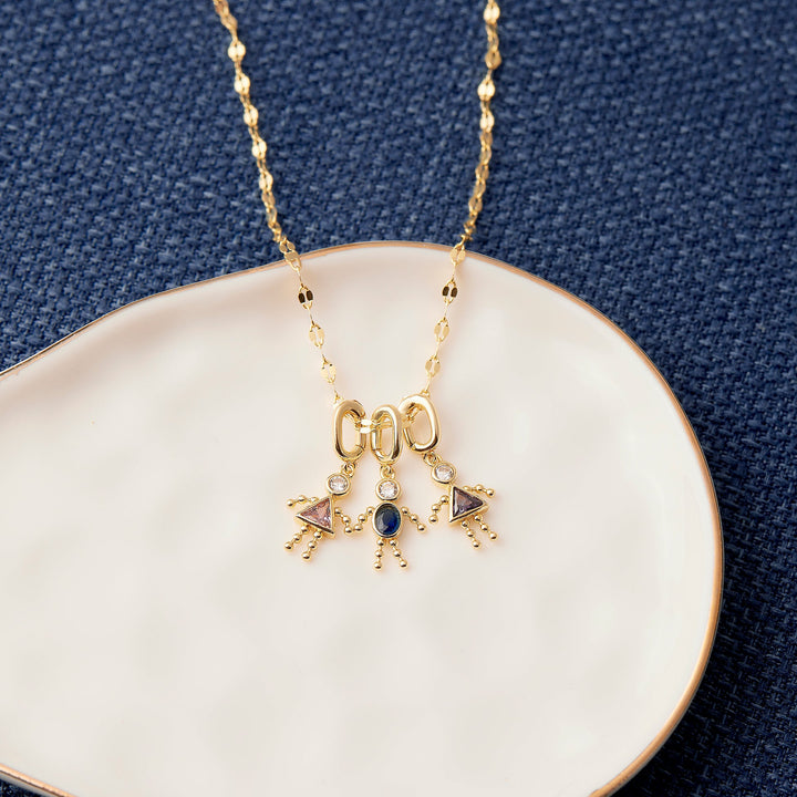 Birthstone child charm necklace with three dangling figure charms set with birthstones, resting on a ceramic dish.