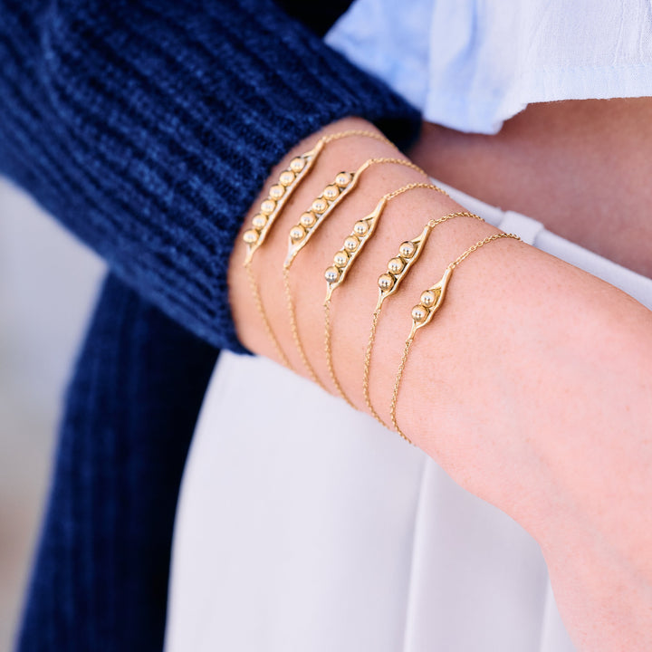 Set of pea pod bracelets with varying pea counts, worn by a model, each with a delicate chain, stacked together on a wrist.