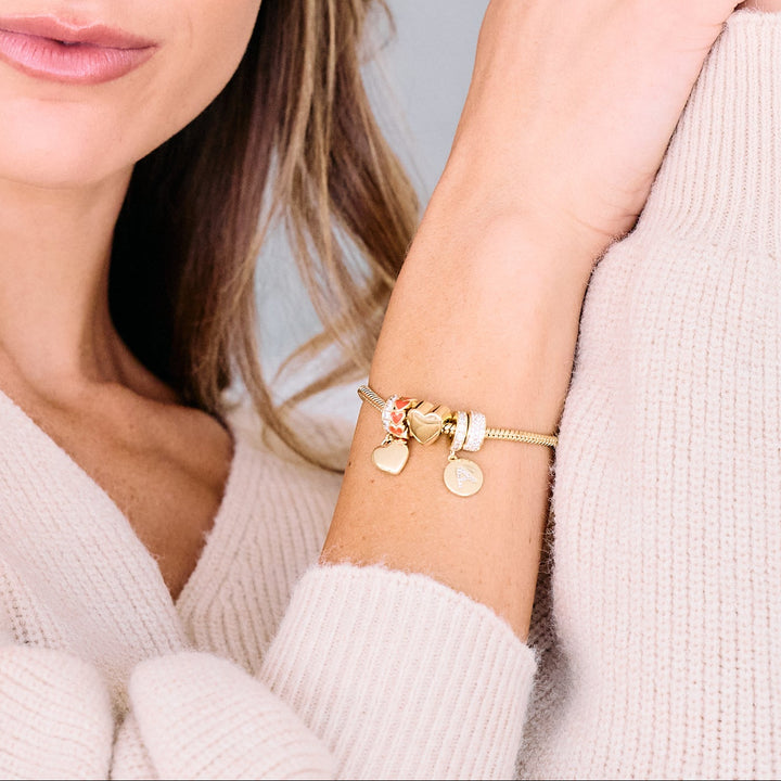 Charm bracelet worn by a model, styled with heart charms, a pavé bead, and an initial disc charm on a ridged bracelet with a round clasp.