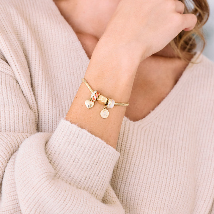 Charm bracelet worn by a model, styled with a heart charm, red enamel bead, pavé ring bead, and a round initial disc charm dangling from a ridged bangle.