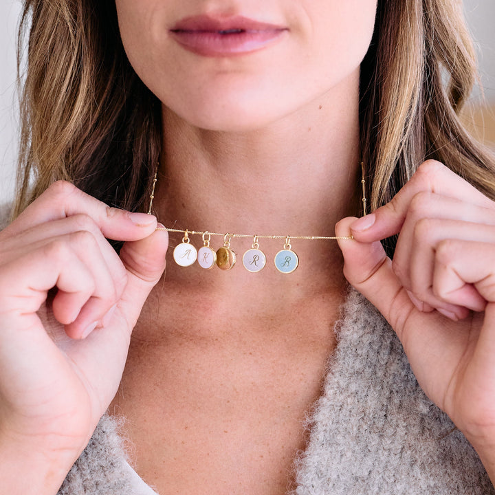 Initial charm necklace with multiple round script-engraved discs in white and blue enamel plus one plain disc, worn by a model as a choker.