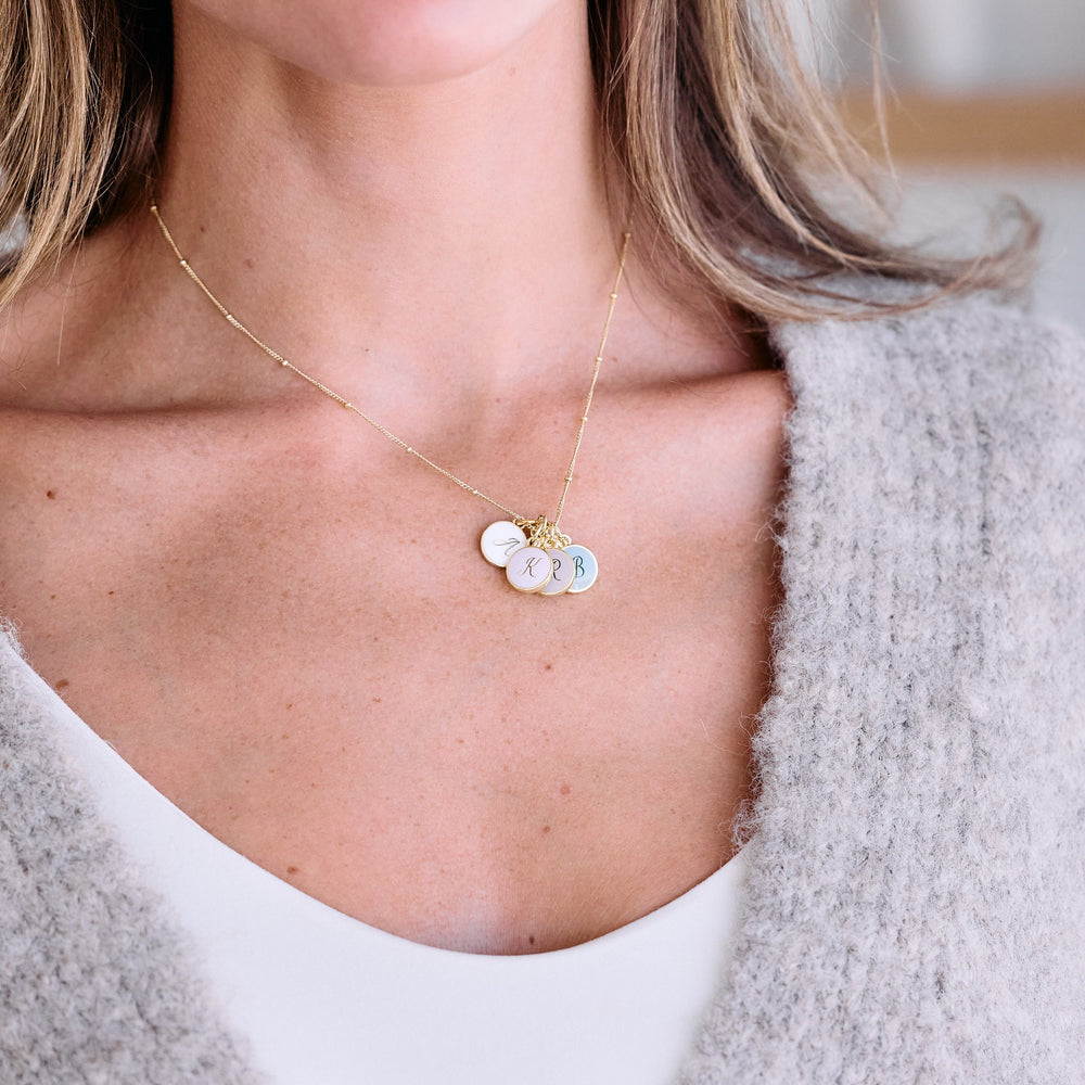 Initial charm necklace with three round script-engraved discs in pastel mother-of-pearl, worn by a model.