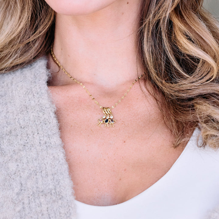 Child charm necklace with multiple birthstone figure pendants on a chain, worn by a model, each charm set with a round and gemstone body detail.