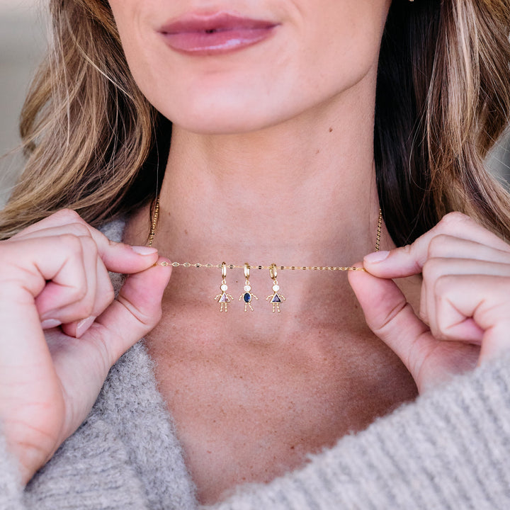 Child charm necklace with three birthstone figure pendants on a chain, worn by a model — each charm depicts a child silhouette with a bezel-set stone.